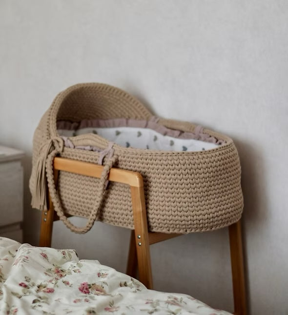 A crocheted moses basket on a wooden stand beside a bed
