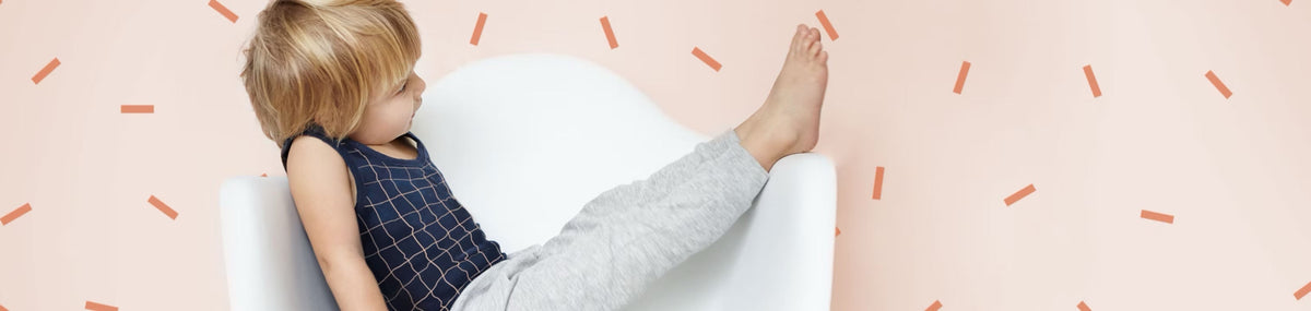 A toddler sitting back in a small white chair against a blush wall